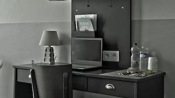 A modern hotel room with a desk, chair, flat-screen TV, laptop, and a small table with a lamp and bottled water.