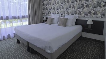 Een hotelkamer met een groot bed, een bekleder met patroon en uitzicht door dunne gordijnen.
