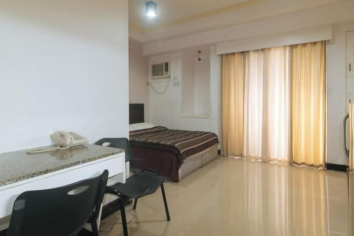 A hotel room with a bed, a desk with a telephone, a chair, and a window with curtains.