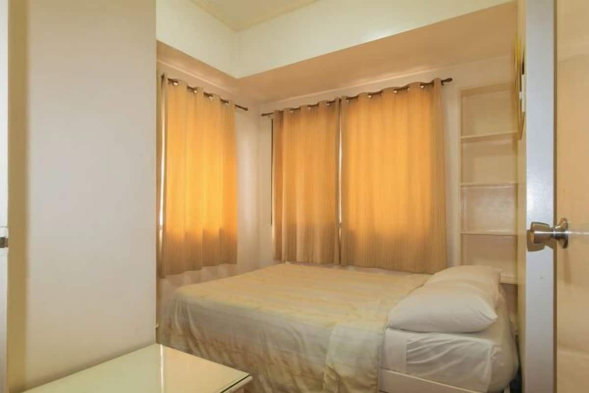 A single bed with a white mattress and pillows, a bedside table, and a wardrobe with a mirror.