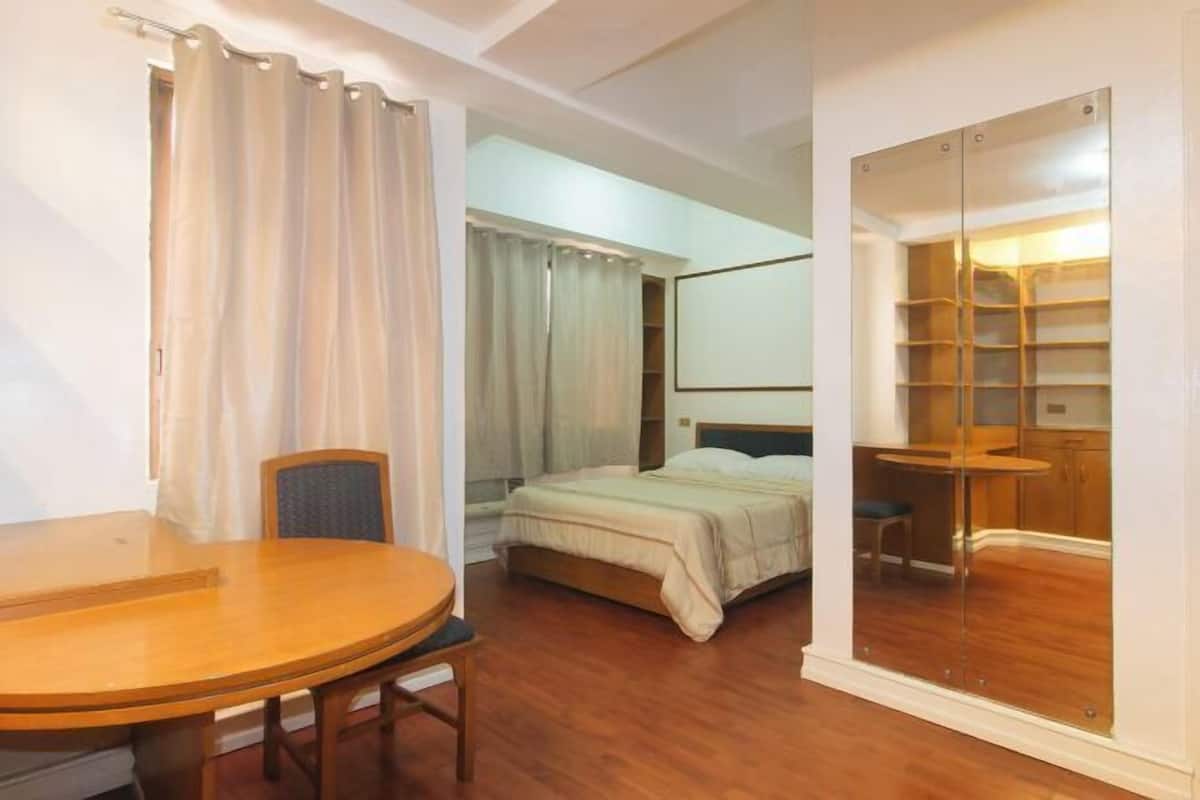 A hotel room with a bed, a wooden table, a chair, a mirror, and a closet with shelves.