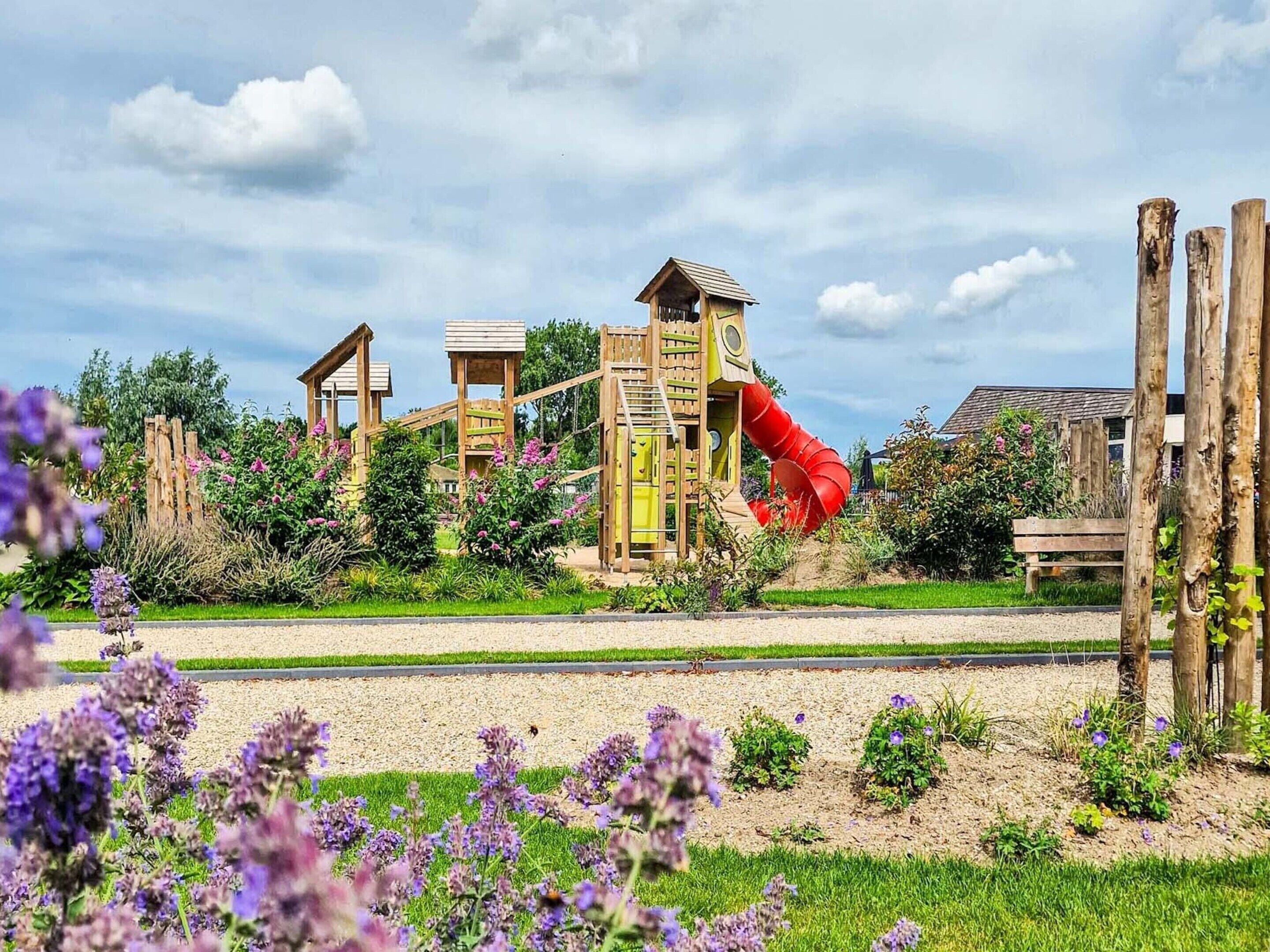 Un terrain de jeux avec un toboggan rouge et des structures d’escalade, entouré d’un jardin où poussent des fleurs violettes.