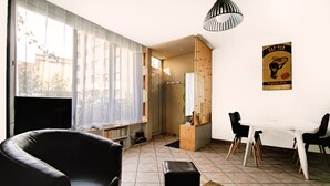 Una sala de estar con un sillón de cuero negro, una mesa de comedor con sillas, una estantería y un ventanal con cortinas translúcidas.