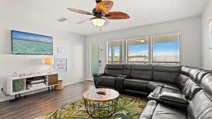 A living room with a black leather sofa, a round coffee table, a ceiling fan, a TV, and a large window with blinds. - Dog Friendly Getaway Steps to Beach & Pool, Coffee Bar, K Bed, Putting Green (Panama City Beach)