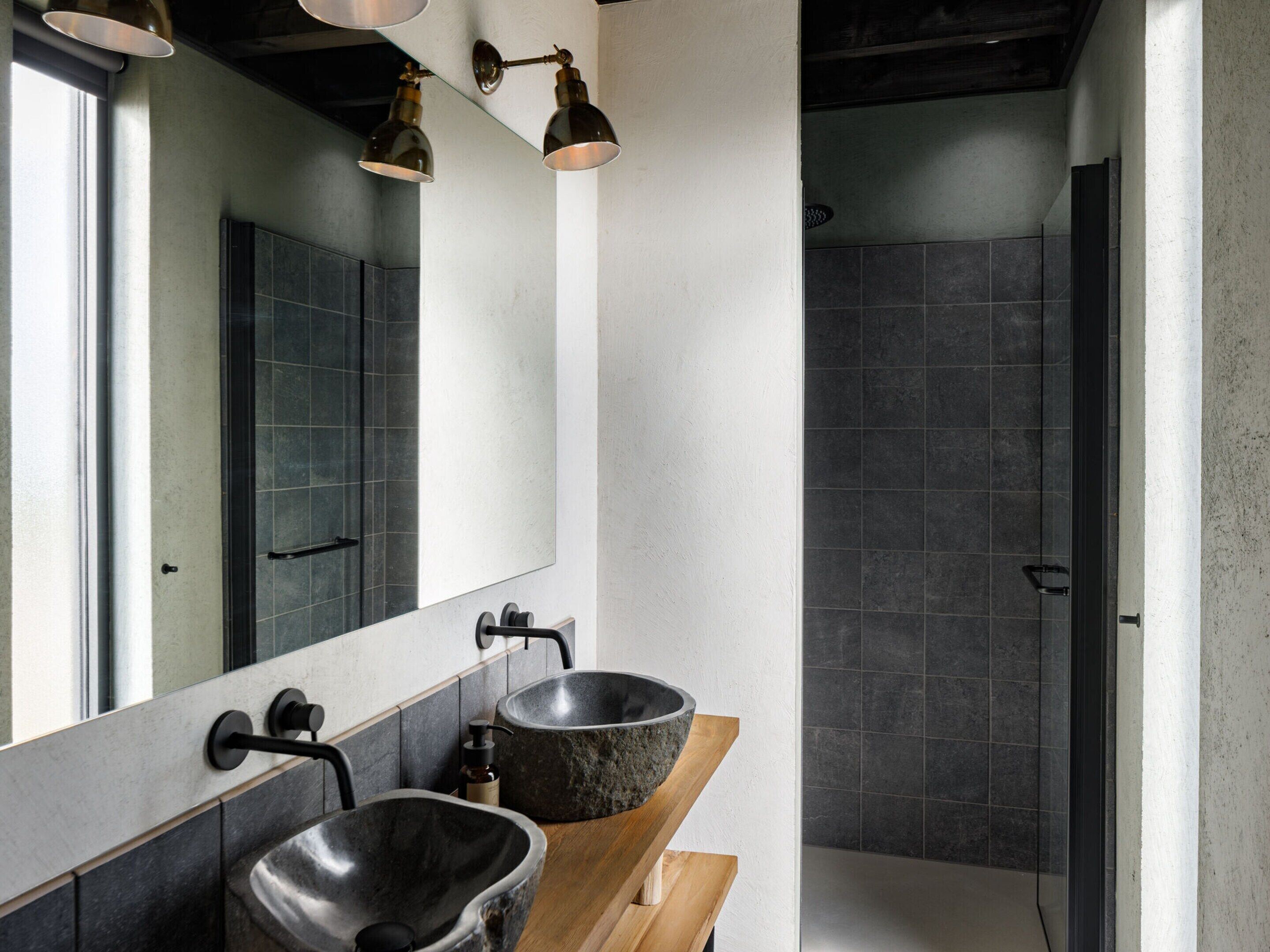 A modern bathroom with two large stone sinks, a wooden countertop, a large mirror, and a shower area with dark tiles.