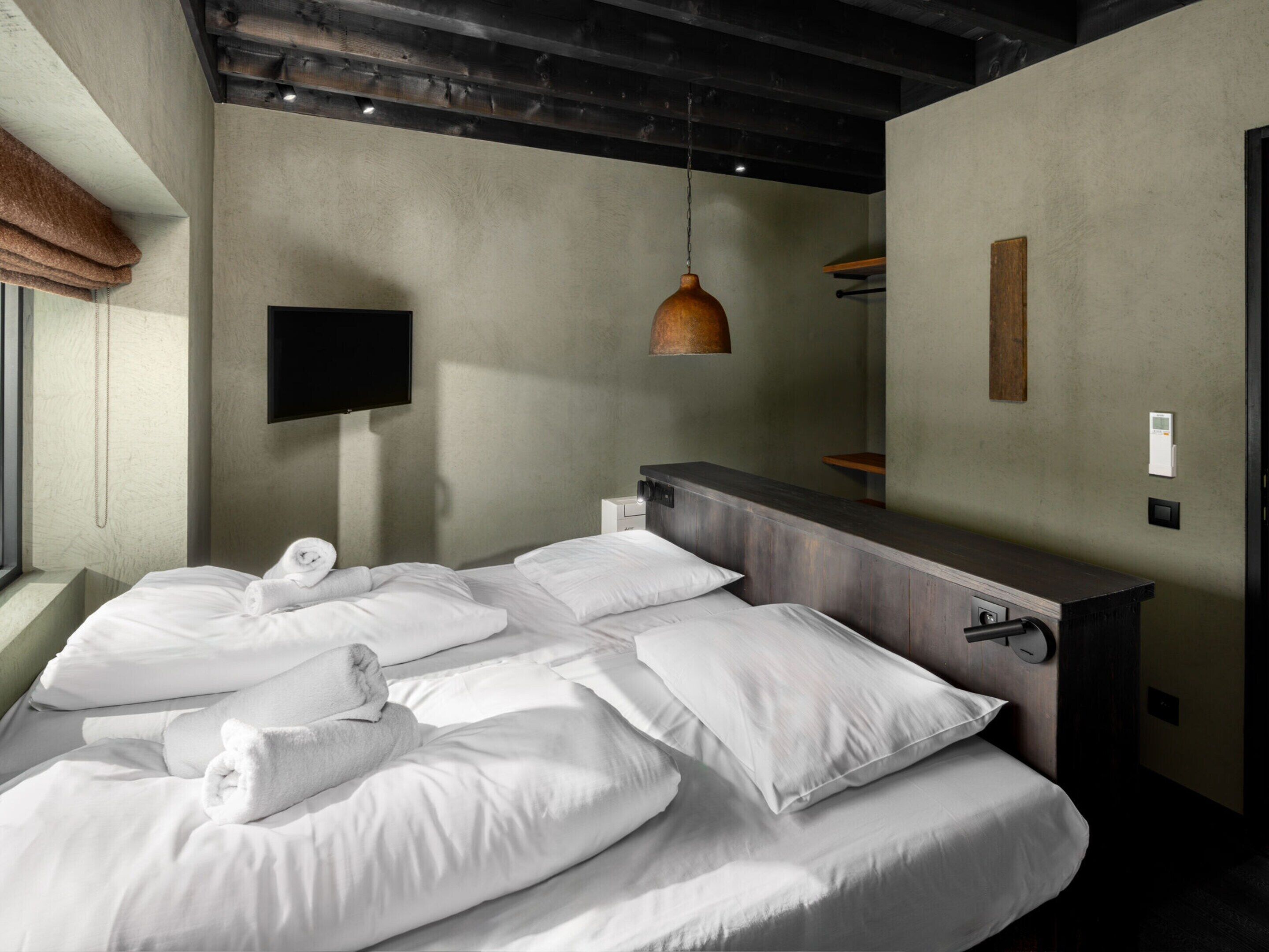 A modern hotel room with a large bed, a wooden bedside table, a mounted TV, and a pendant light.