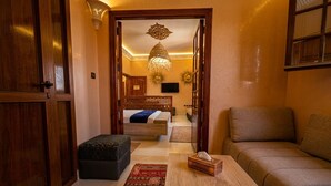 Une pièce avec une porte en bois, un canapé, une petite table, un lit avec une tête de lit, un luminaire suspendu et un téléviseur fixé au mur.