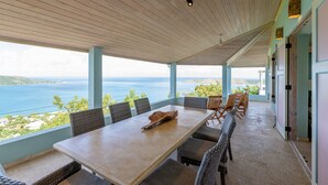 Ein überdachter Essbereich im Freien mit Tisch und Stühlen, mit Blick auf eine schöne Landschaft aus Meer und Hügeln.