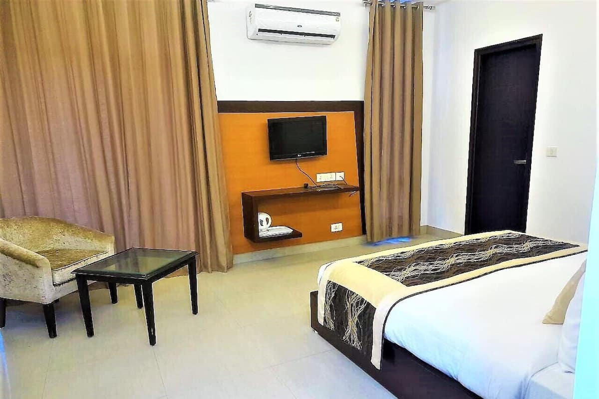 A hotel room with a bed, a chair, a small table, a TV, and an air conditioning unit.