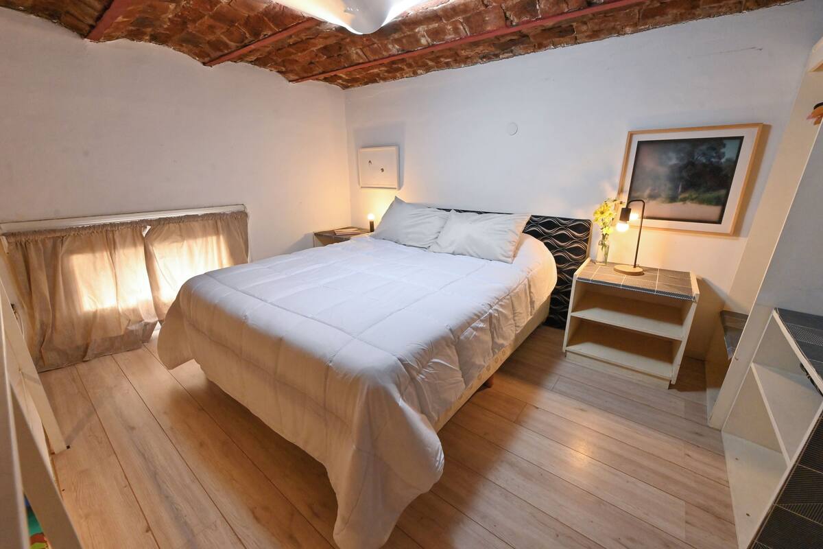 Ein Schlafzimmer mit einem großen Bett, Holzboden, einer Ziegeldecken und einem Nachttisch mit Lampe, sowie einem gerahmten Bild an der Wand.