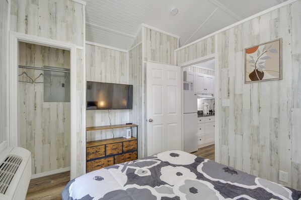 A bedroom with a bed, a dresser, a TV, and a door leading to a kitchen.