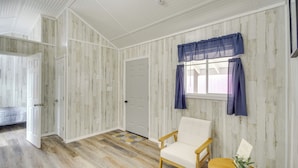 A room with wooden panel walls, a white door, a window with blue curtains, a white chair, and a small table with a plant. - Pet-Friendly Picayune Tiny Home Near Space Center! (Picayune)