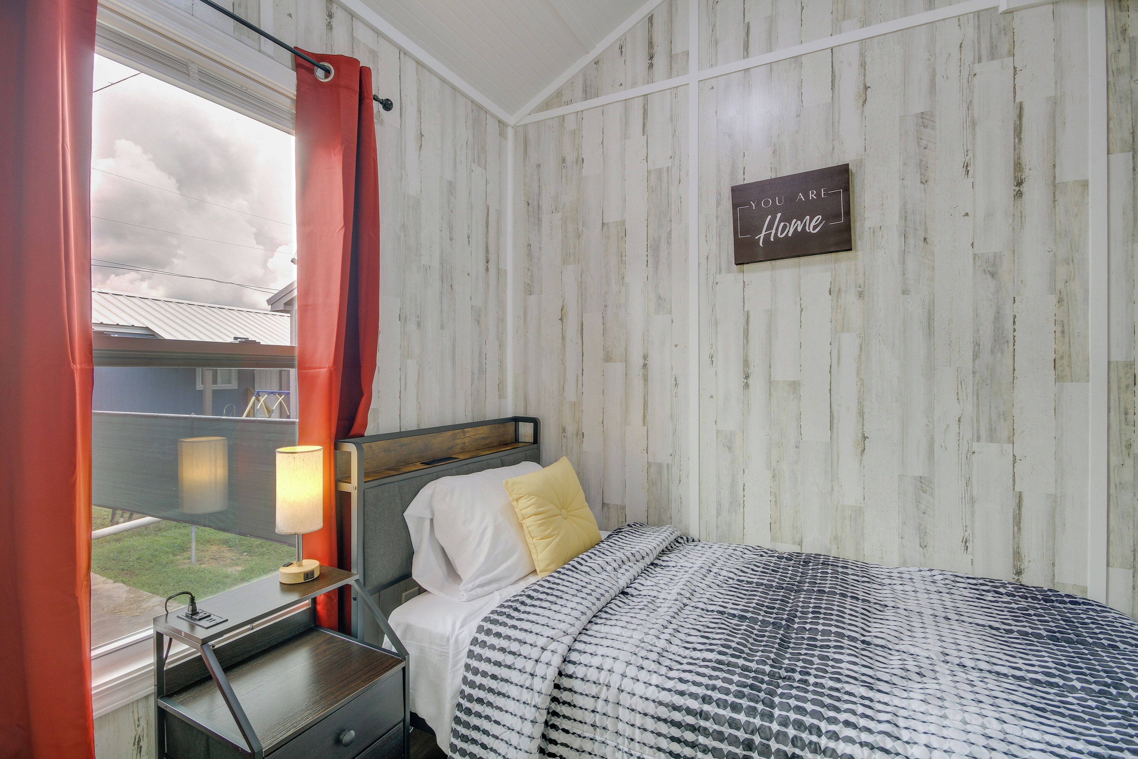 Ein Schlafzimmer mit Bett, Nachttisch mit Lampe, Fenster mit Vorhängen und einem Schild an der Wand mit der Aufschrift 'You are Home'.