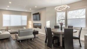 Un salon moderne avec un canapé gris, une table à manger avec des chaises, un téléviseur à écran plat fixé au mur et une grande fenêtre donnant sur les arbres.