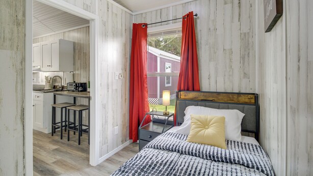 A bedroom with a bed, a nightstand, a desk, and a chair. There is a window with red curtains.