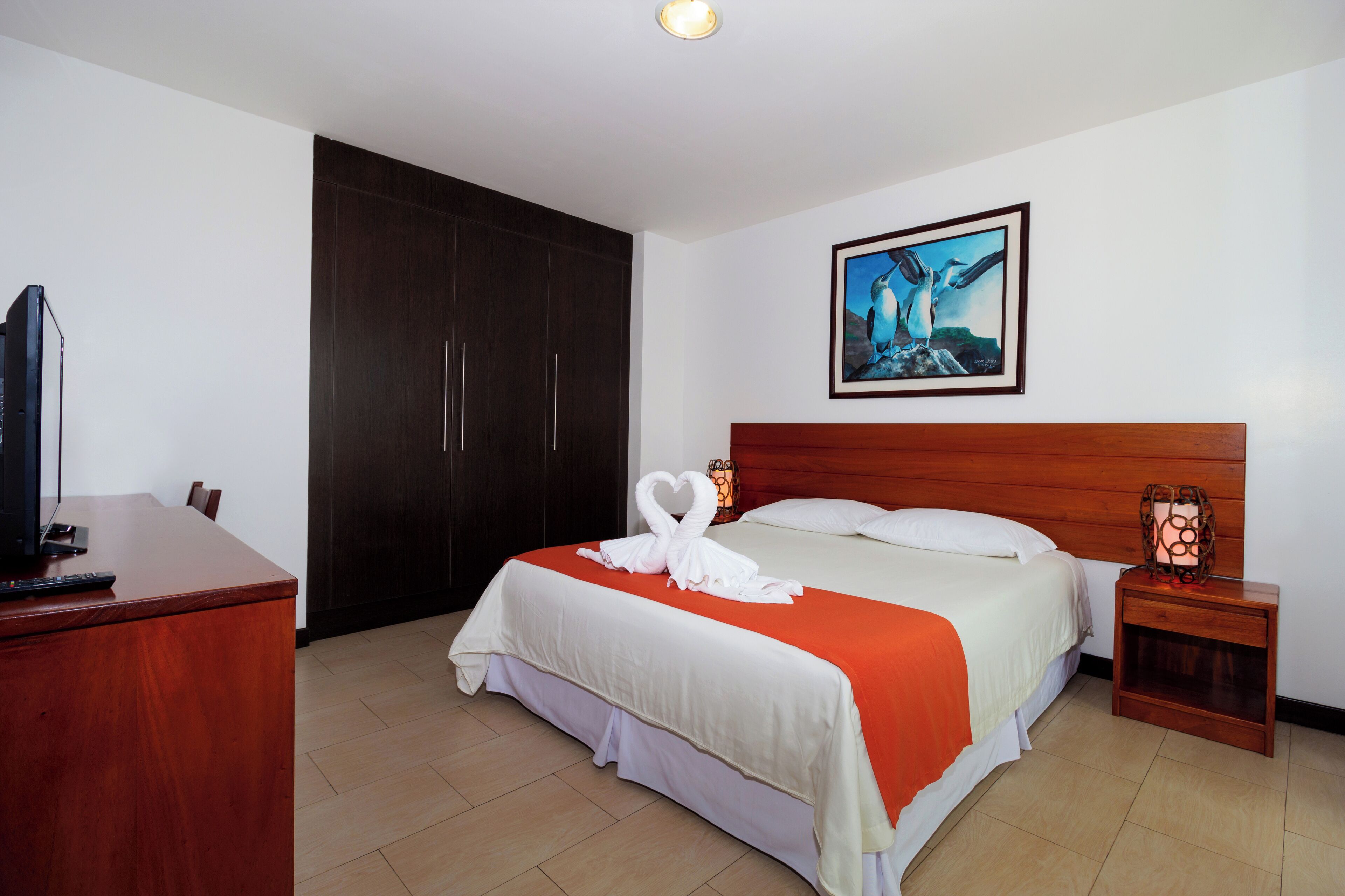 A hotel room with a bed, a desk, a TV, a wardrobe, and a painting on the wall.