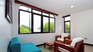 Una sala de estar con un sofá color turquesa, sillones de madera, una mesa de centro y ventanas amplias con vistas a zonas verdes.
