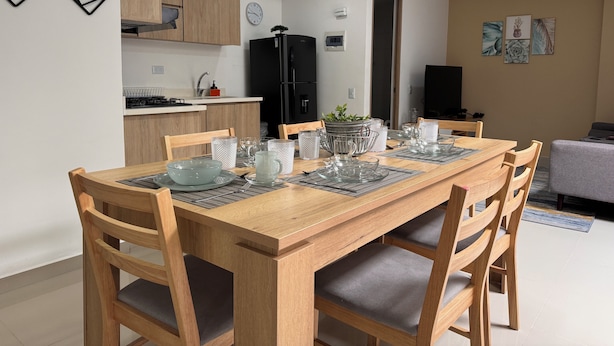 A modern dining area with a wooden table set for four, surrounded by wooden chairs with grey cushions. A kitchen with wooden cabinets and a black refrigerator is visible in the background. - Exclusivo Apartamento en el Poblado by Hoost 705 (Medellín)