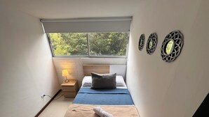 A bedroom with a bed, a nightstand, a lamp, and a window with a view of trees. - Exclusivo Apartamento en el Poblado by Hoost 705 (Medellín)