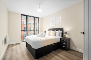 Un dormitorio moderno con una cama grande, mesitas de noche y vista a un edificio de ladrillo a través de la ventana.