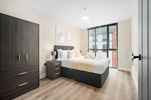 Une chambre moderne avec un lit, une armoire, une table de chevet et une vue sur un immeuble par la fenêtre.