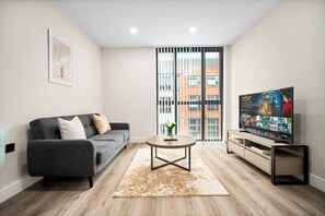 A modern living room with a grey sofa, a coffee table, a TV on a stand, and a large window with a view of buildings. - Stylish 2 Bed Apartment - Smart TV - WIFI -  Jewellery Quarter (Birmingham)