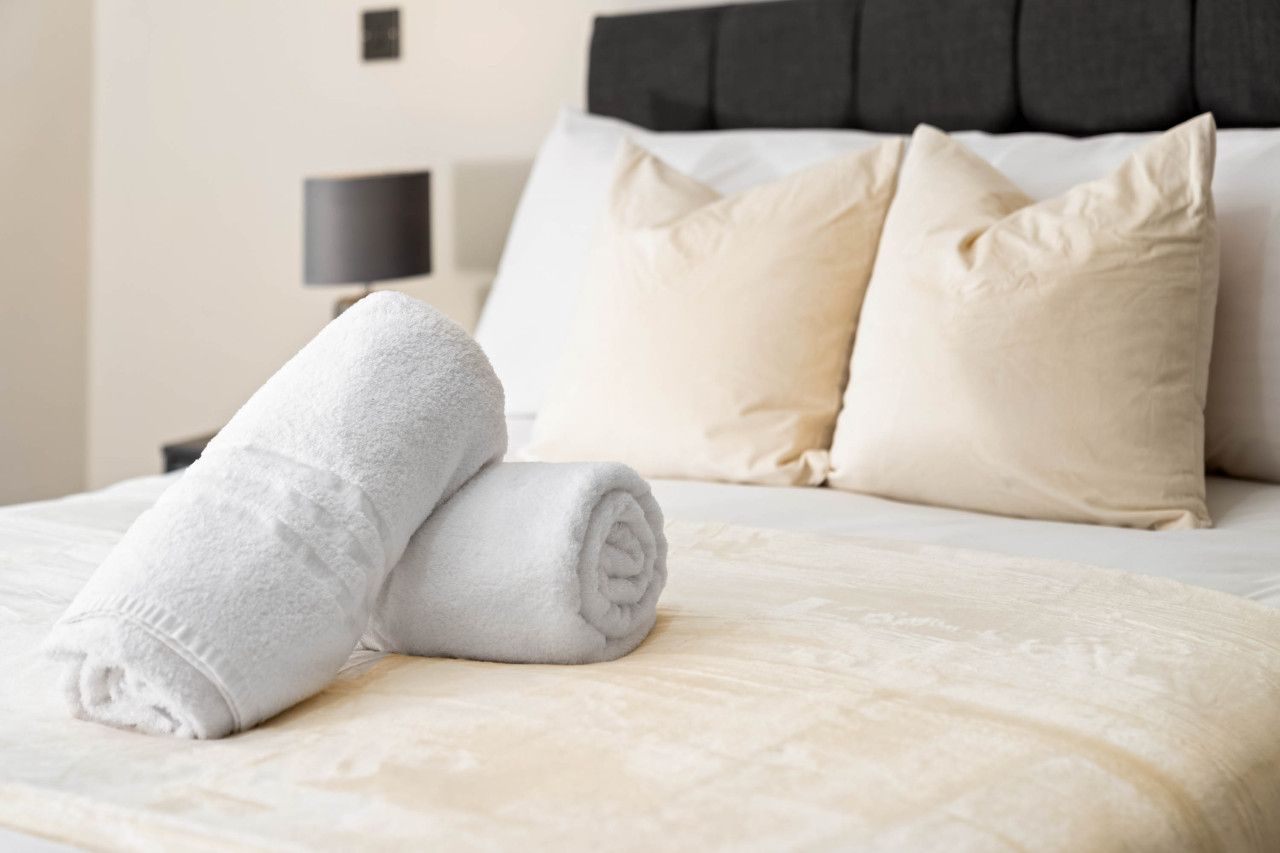 Ein ordentlich gemachtes Bett mit einem aufgerollten weißen Handtuch, einem dunklen Kopfteil und einer Nachttischlampe.