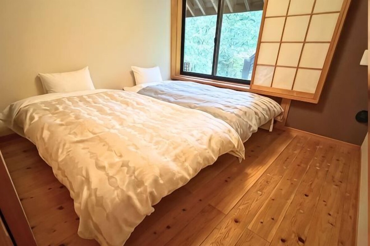 A bedroom with a wooden floor, a bed with white bedding, and a window with a view of greenery.