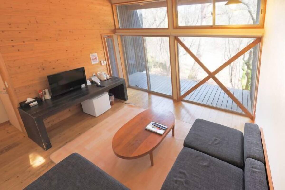 Un salon moderne avec un plancher en bois, une table basse en bois foncé, un canapé gris, un téléviseur à écran plat posé sur un meuble en bois et de grandes fenêtres donnant sur l’extérieur.