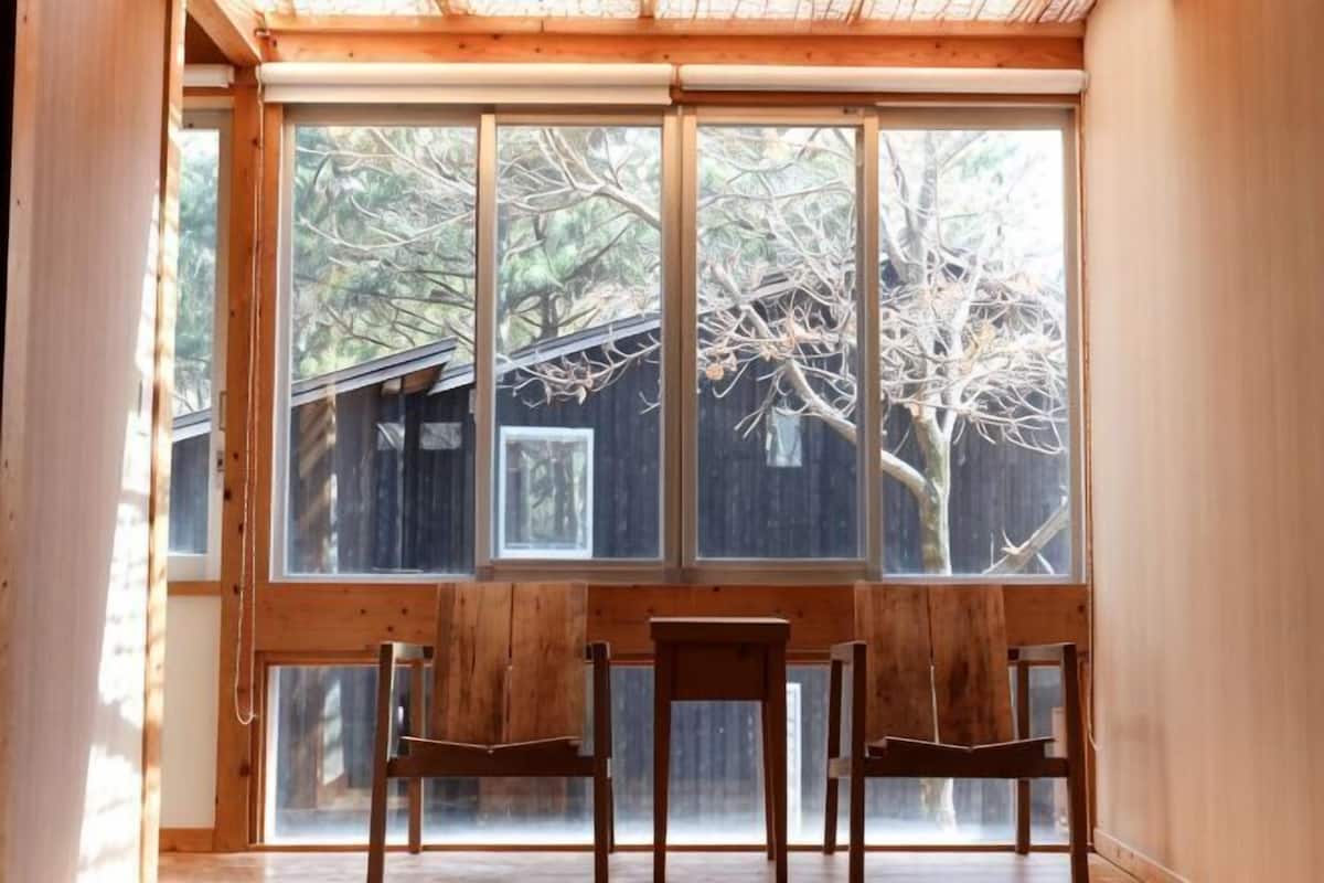 Une pièce dotée d’une grande fenêtre, de chaises en bois et offrant une vue sur des arbres et une structure en bois à l’extérieur.