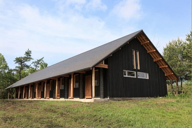 A large black barn with a sloped roof and wooden beams, surrounded by grass and trees.