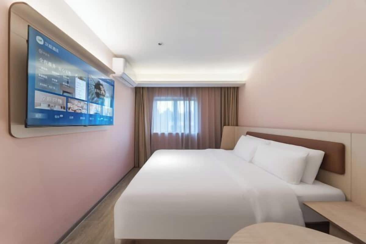 A hotel room with a large flat-screen TV mounted on the wall, a bed with white linens, a wooden headboard, and a window with curtains.