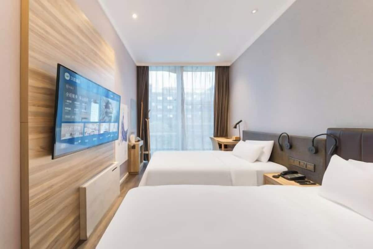 Une chambre d’hôtel avec deux lits, un téléviseur à écran plat fixé au mur, un bureau et une fenêtre avec des rideaux.