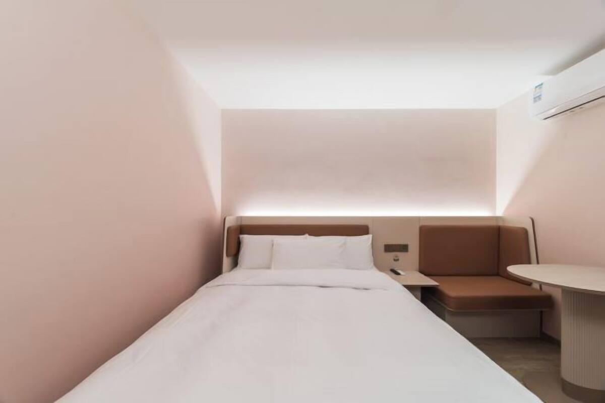 A small, modern hotel room with a single bed, a nightstand, a seating area, and an air conditioning unit.