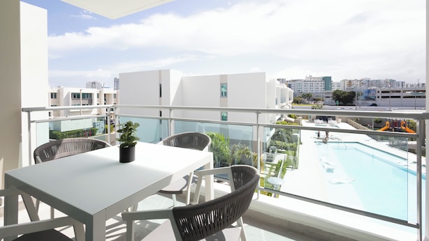 Un balcón con una mesa blanca y sillas de mimbre, con vista a una piscina y al horizonte de la ciudad.