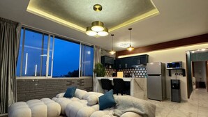 A modern living room with a sectional sofa, a glass coffee table, and a kitchen area with a refrigerator, microwave, and bar stools.