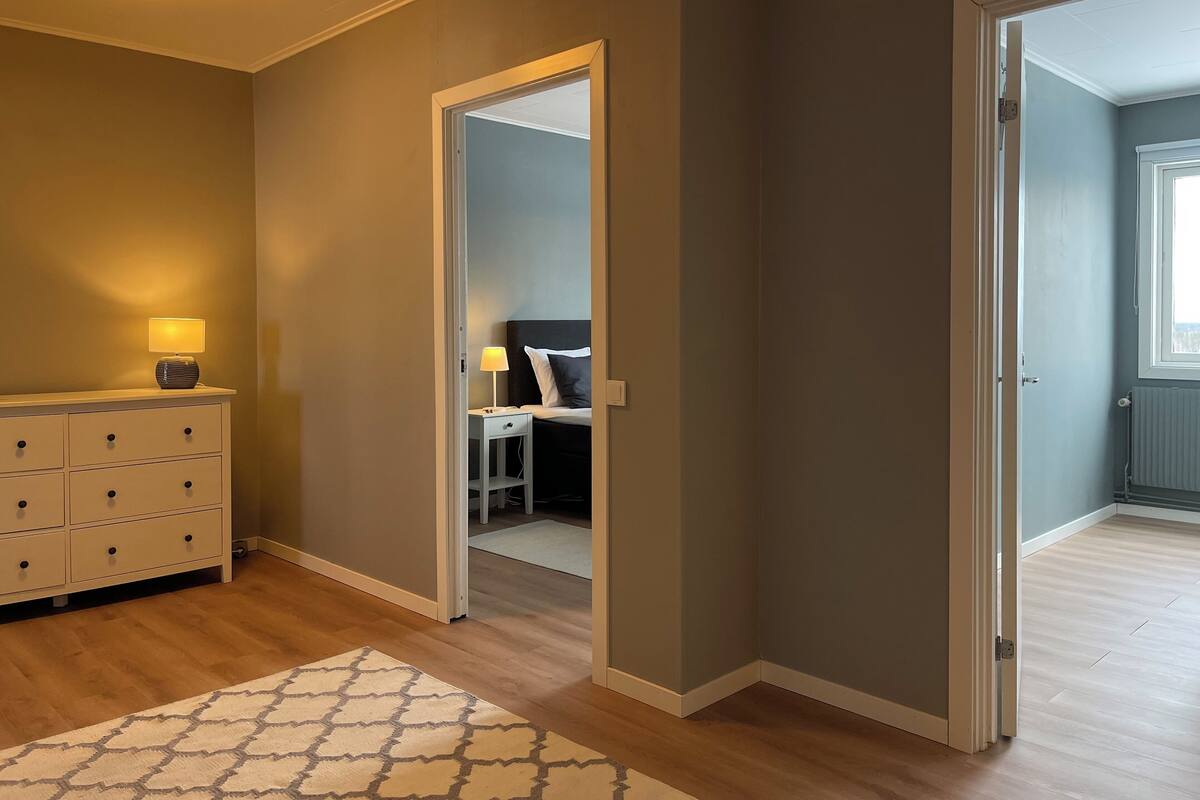 A hotel room with a wooden floor, a white dresser with drawers, a patterned rug, and a door leading to another room.