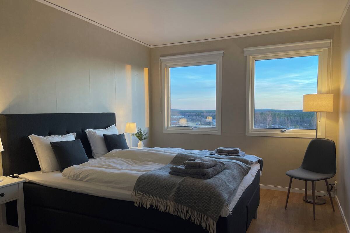 A modern hotel room with a large bed, two windows with a view, a bedside table, and a chair.
