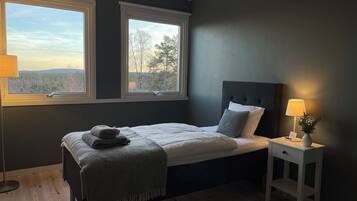 A bedroom with a bed, two windows, a bedside table with a lamp, and a view of trees and hills.