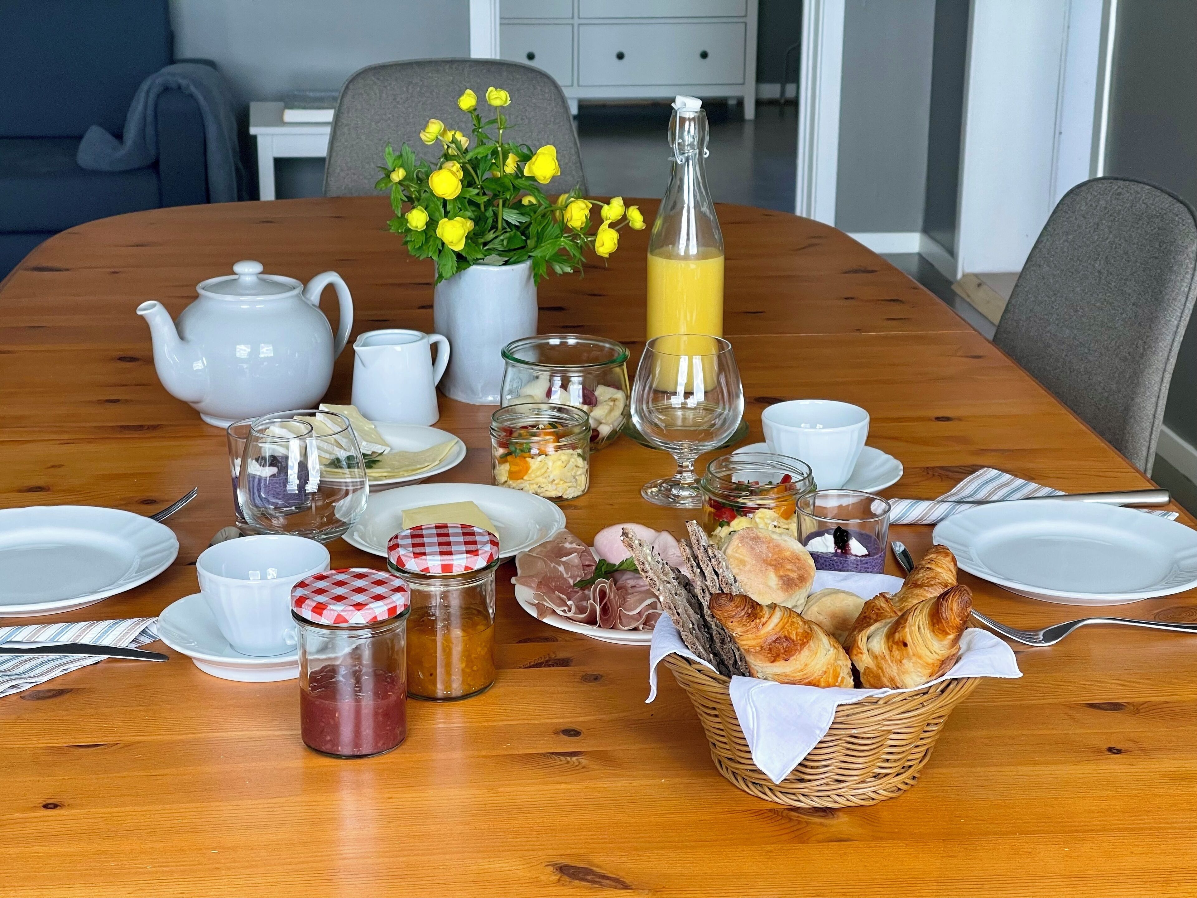 Una mesa de desayuno dispuesta con platos, vasos, una tetera, una jarra, una botella de jugo de naranja, un florero con flores amarillas y una cesta de cruasanes.