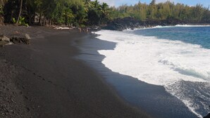 A black sand beach with waves crashing onto the shore, surrounded by palm trees and a rocky cliff. - Kehena Black Sand Beach House - ocean view - swim spa (Pahoa)
