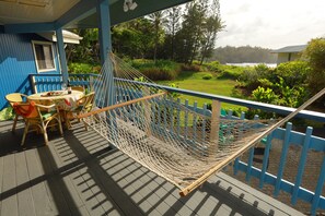 A hammock on a wooden deck with a table and chairs, overlooking a lush garden and a body of water.