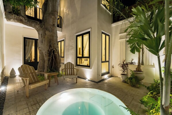Un espacio exterior bien iluminado con un jacuzzi, sillas de madera y plantas en macetas.
