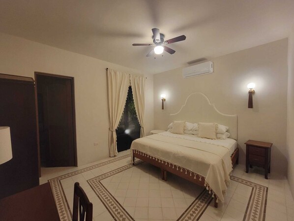 A bedroom with a bed, a ceiling fan, an air conditioning unit, a nightstand, and a window with curtains. - Luxury Historic 5BR Boutique hotel (Mérida)