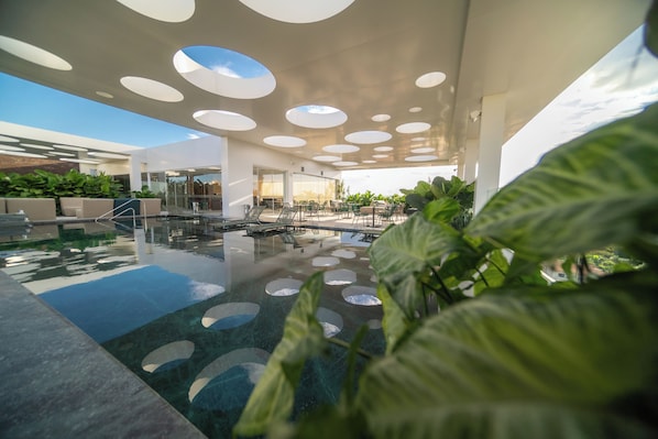 A modern indoor pool area with circular skylights, lounge seating, and a glass door entrance. - Mona Hotel (Teresina)