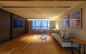 A modern living room with a wooden floor, a sofa, a coffee table, and two chairs. There is a TV mounted on the wall and a window with curtains. - Mona Hotel (Teresina)