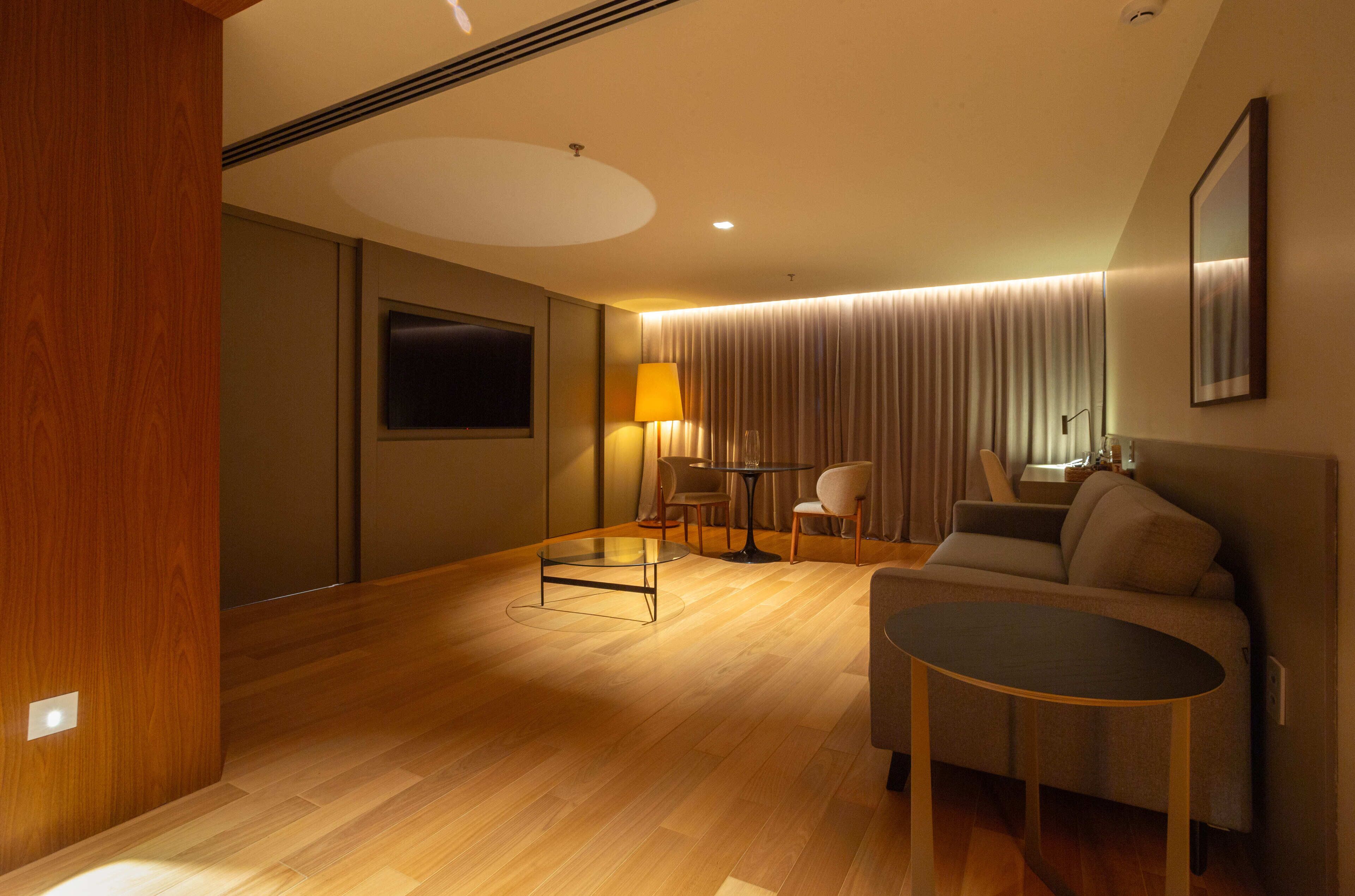 A modern hotel room with a wooden floor, a sofa, a round table, a TV, and a lamp.