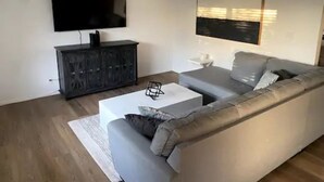 Una sala moderna con un sofá gris, una mesa de centro blanca, un mueble negro y un televisor de pantalla plana montado en la pared.
