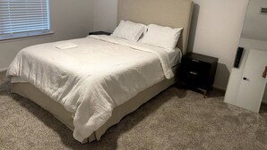 Una cama bien hecha con edredón blanco y almohadas, una mesita de noche con lámpara y un armario adosado a la pared.