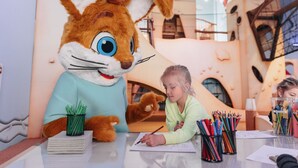 Une personne déguisée en lapin est assise à une table, devant du matériel de dessin, dans une pièce dotée d’une architecture moderne et d’une grande fenêtre.